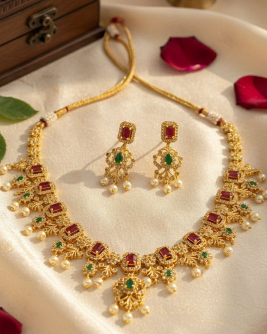 Gold necklace and earrings with red and green stones on a soft surface with leaves and petals.